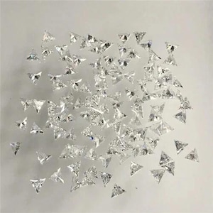 Könnte beinhalten: Eine Ansammlung kleiner, klarer, dreieckig geschliffener Edelsteine. Die Edelsteine sind auf einer hellen Oberfläche verstreut, reflektieren das Licht und erzeugen einen funkelnden Effekt. Die dreieckige Form ist durchgängig.
