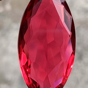 May include: A faceted, marquise-cut gemstone in a deep, vibrant red hue. The gem's surface is a complex arrangement of polished facets, reflecting light and creating a sparkling effect. The elongated shape and rich colour suggest it could be a ruby or garnet.