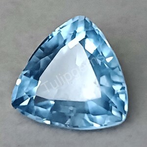 Puede incluir: Una gema triangular facetada de color azul claro. La gema tiene una superficie pulida y un aspecto claro, con el texto "Tulipgems" visible. La gema está sobre un fondo gris neutro.
