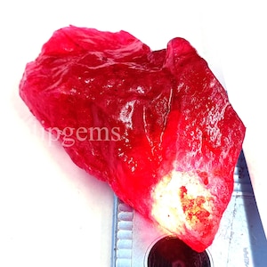 May include: A raw, uncut ruby gemstone with a vibrant red hue. The rough stone has a textured surface and a lighter, almost white area on one side. The word "pingems" is partially visible on the stone.