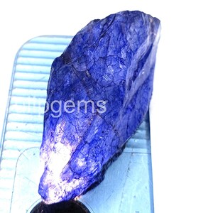 May include: A raw, uncut, deep blue gemstone with a rough, textured surface. The gemstone is displayed on a light blue surface. The word "gems" is visible on the surface.