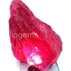 May include: A large, rough-cut ruby gemstone with a vibrant red hue. The raw mineral displays a textured surface and a natural, irregular shape. The image includes the text "lipgems".