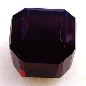 May include: A faceted, dark purple gemstone with an emerald cut. The gem has a rectangular shape with beveled corners and a polished surface. The color is a deep, rich shade of purple, with a hint of red visible on one edge.