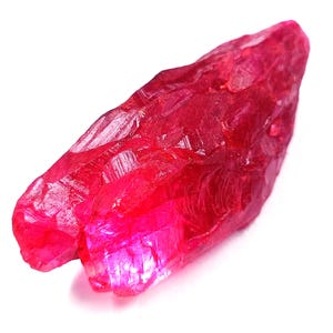 Puede incluir: Una gema de rubí en bruto sin cortar. El rubí es de un color rojo intenso y vibrante con una estructura cristalina. La gema es alargada y tiene una textura rugosa y natural, mostrando su estado sin refinar. El fondo es blanco.