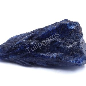 Puede incluir: Una gema de zafiro en bruto, sin cortar, en tonos azul oscuro. La piedra en bruto tiene una superficie texturizada y una forma irregular. El texto "Tulipgems" es visible.