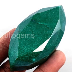 Puede incluir: Una gema facetada de color verde esmeralda con talla marquesa. La gema se sostiene en una mano y la superficie refleja la luz, mostrando sus múltiples facetas. El texto "tulipgems" es visible.