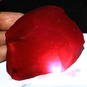 May include: A large, raw, uncut gemstone with a deep, vibrant red color. The stone has an irregular, organic shape with a translucent quality that allows light to pass through, creating a bright, glowing pinkish-red highlight on one side.