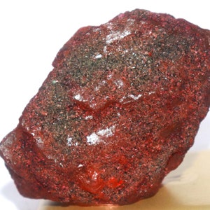 May include: A rough, irregularly shaped mineral specimen with a deep red hue, speckled with darker, possibly metallic, inclusions. The surface has a slightly glossy appearance, suggesting a polished or naturally smooth texture. The overall form is organic and natural.