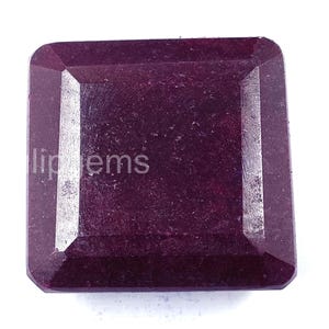 Puede incluir: Una gema roja oscura de corte cuadrado con una superficie pulida. La gema tiene forma rectangular con bordes biselados y el texto "lip gems" es visible en el lado izquierdo. La piedra está sobre un fondo blanco.