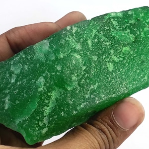 Può includere: Una gemma verde vibrante, tagliata grezza, con una superficie strutturata. Il minerale grezzo è tenuto in una mano, mostrando la sua forma e il suo colore naturali e irregolari. La pietra ha un aspetto cristallino.
