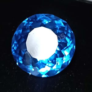 Puede incluir: Una gema redonda tallada en brillante, de color azul profundo. La gema presenta un centro blanco brillante y está colocada sobre un fondo oscuro. Las facetas crean un juego de luces.