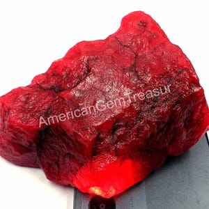 May include: A large, rough-cut ruby gemstone with a deep red color. The surface has a textured appearance, and the text "American Gem Treasur" is visible on the surface. The gemstone is displayed on a light gray surface.
