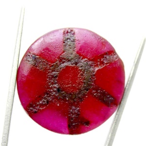 May include: A round, polished ruby gemstone with a deep red hue and a dark, star-like inclusion pattern. The gemstone is held by silver tweezers against a white background, showcasing its vibrant color and unique internal features.