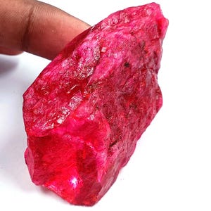 May include: A rough, uncut ruby gemstone with a vibrant, deep red color. The raw mineral displays a textured surface and irregular shape, showcasing its natural form. The gemstone is held against a white background.