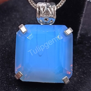 May include: A square, light blue pendant with a silver-colored setting and chain. The pendant has a rectangular shape with a polished surface. The text "Tulipgems" is visible on the pendant.