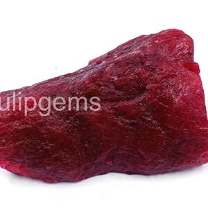 Pode incluir: Uma gema de rubi bruta e não lapidada, de cor vermelha escura. A superfície é texturizada e irregular, com estruturas cristalinas visíveis. A palavra "ulipgems" está impressa na superfície da gema.