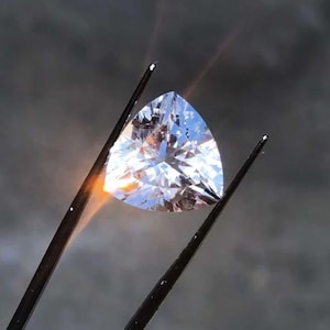 Könnte beinhalten: Ein klarer, dreieckig geschliffener Edelstein wird von einer Pinzette vor grauem Hintergrund gehalten. Der Edelstein funkelt und reflektiert das Licht. Die Pinzette ist schwarz. Das Thema ist Schmuck.