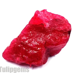 May include: A rough, uncut ruby gemstone with a vibrant red hue. The raw mineral has a textured surface and irregular shape, showcasing its natural form. The image includes the text "Tulipgems" at the bottom.