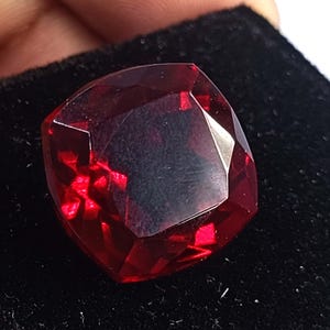May include: A cushion-cut gemstone with a deep, vibrant red color and multiple facets that reflect light. The stone has a square shape with rounded corners and a polished, transparent surface, displaying internal light reflections throughout.