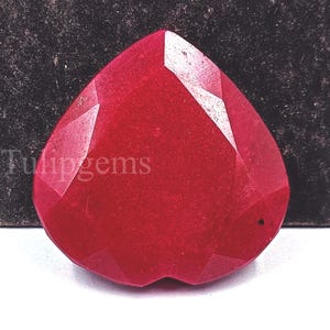 Puede incluir: Una gema de rubí facetada en forma de corazón. La gema es de color rojo intenso y saturado, con múltiples facetas que captan la luz. El texto "Tulipgems" es visible en el lado izquierdo.