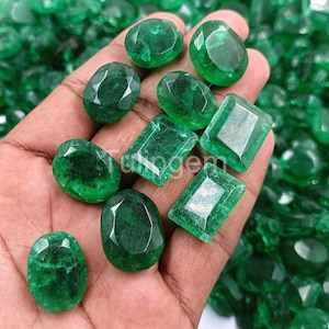 Op de afbeelding: Een verzameling gefacetteerde smaragd edelstenen in verschillende vormen, waaronder ovale en rechthoekige slijpsels. De edelstenen hebben een levendige groene kleur en worden in een hand gehouden, met meer smaragden zichtbaar op de achtergrond.