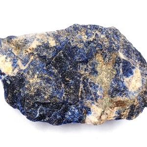 May include: A rough-hewn, natural sodalite stone. The rock features a deep blue color with streaks of white and tan throughout. The stone has a rugged texture and irregular shape, showcasing its raw, unpolished state.