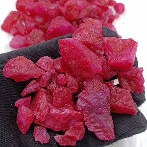 May include: A collection of rough, uncut ruby gemstones. The raw rubies are a deep, vibrant red color, with a slightly textured surface. The gemstones are in various sizes and shapes, with a natural, unpolished appearance.