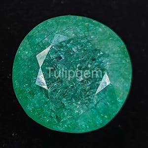 Puede incluir: Una gema de esmeralda redonda y facetada, de color verde intenso. La gema tiene una superficie pulida con inclusiones internas visibles y el texto "Tulipgem" es visible. La gema está sobre un fondo negro.