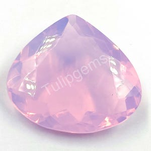 Puede incluir: Una gema facetada en forma de corazón, en tonos rosa y lavanda. La gema tiene una superficie pulida con múltiples facetas que reflejan la luz. El texto "Tulipgems" está impreso en la superficie de la gema.