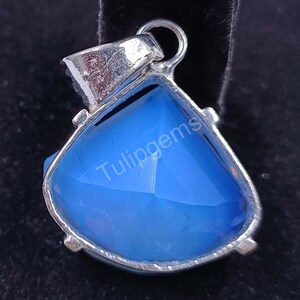 May include: A silver-toned pendant with a large, triangular, faceted blue gemstone. The pendant has a loop at the top for a chain and the text "Tulipgems" is visible on the gemstone.