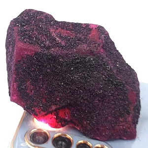 Peut inclure: Une pierre précieuse de rubis brute, d'un rouge profond, partiellement recouverte d'une texture sombre et granuleuse. La surface de la pierre reflète la lumière, révélant son état naturel et non poli. Un exemple vibrant de spécimen minéral brut.
