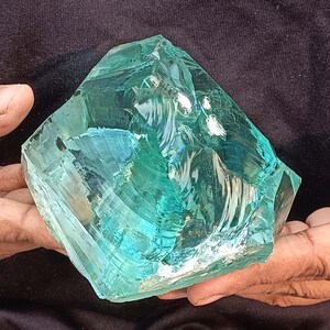 May include: A large, irregularly shaped piece of translucent, teal-coloured glass. The glass has a rough, faceted surface with internal reflections, held in a person's hands. The overall appearance suggests a natural, raw gemstone or decorative element.