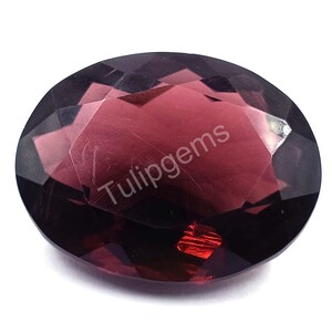 May include: Oval-cut, faceted gemstone with a deep burgundy color. The gem has a polished surface with multiple facets that reflect light. The text "Tulipgems" is printed on the surface. The gemstone is set against a white background.