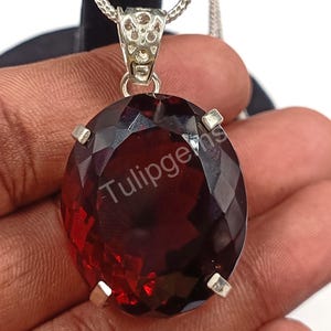 Op de afbeelding: Een dieprode, ovaal geslepen edelsteen hanger met een zilveren zetting en ketting. De edelsteen is gefacetteerd. De tekst "Tulipgems" is zichtbaar op de edelsteen.