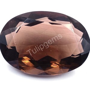 May include: Oval-cut smoky quartz gemstone with a faceted surface. The gem is a translucent brown color with a polished finish. The text "Tulipgems" is visible on the surface of the gemstone.