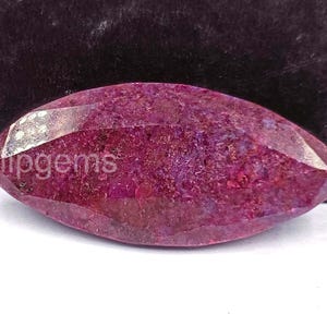 Puede incluir: Una gema facetada de forma ovalada con un tono magenta intenso. La piedra tiene una superficie pulida con múltiples facetas, que reflejan la luz. El texto "Tulipgems" es visible en el lateral de la piedra. La gema está sobre un fondo oscuro.