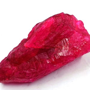 May include: A raw, uncut ruby gemstone with a deep red hue. The rough surface of the stone displays natural textures and facets, reflecting light. The ruby is a vibrant, eye-catching specimen, perfect for collectors or jewelry making.