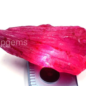 Pode incluir: Uma gema de rubi bruta, não lapidada, com um tom rosa-avermelhado vibrante. A pedra bruta tem uma superfície texturizada e é iluminada por baixo, revelando sua estrutura cristalina natural. O texto "tulipgems" é visível na pedra.