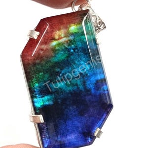 Può includere: Un ciondolo con pietra preziosa multicolore con una cauzione in argento. Il ciondolo ha una forma rettangolare con un gradiente di colori, tra cui rosso, verde e blu. Il testo "Tulipgems" è visibile.