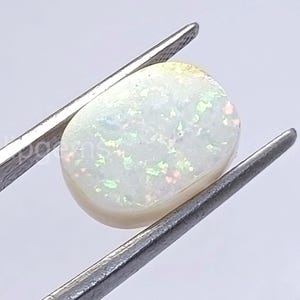May include: A polished, oval-shaped opal gemstone, held by silver tweezers. The opal has a milky white base color with iridescent flashes of green, pink, and blue throughout its surface. The gemstone is smooth and rounded.