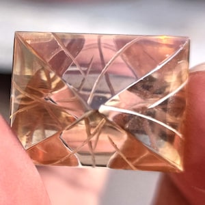 May include: A rectangular, faceted gemstone with a warm, peach colour. The gem features a complex pattern of intersecting lines, creating a geometric design. The light reflects off the facets, giving it a sparkling appearance. The gemstone is held between fingers.