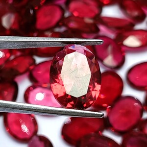 May include: A faceted, oval-cut red gemstone is held by tweezers, with a scattering of similar gems in the background. The gems have a brilliant, transparent appearance, reflecting light and showcasing their cut.