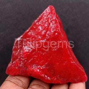 Può includere: Una gemma rosso vivo, grezza, con una consistenza cristallina. La pietra ha una forma triangolare e il testo "Tidipgems" è visibile. La gemma è tenuta in mano su sfondo scuro.