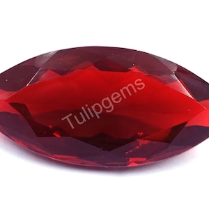 May include: A faceted, marquise-cut gemstone with a deep red hue. The gem has a pointed oval shape and multiple facets that catch the light. The text "Tulipgems" is visible on the surface of the gem.