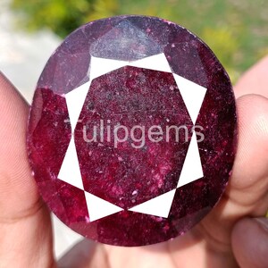 Pode incluir: Uma grande pedra preciosa de rubi de corte oval com um tom vermelho profundo. A gema apresenta um proeminente design geométrico branco no centro, com a palavra "tulipgems" impressa na superfície. A gema é segurada em uma mão.