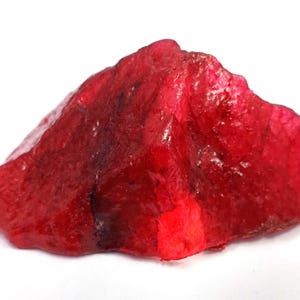 May include: A raw, uncut ruby gemstone in a deep red colour. The natural stone has an irregular shape and a textured surface, showing its raw, unpolished state. The ruby is set against a plain white background.