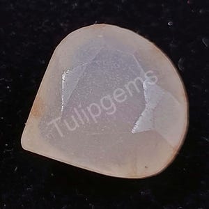 Puede incluir: Una gema translúcida en forma de pera, de color melocotón pálido. La gema tiene una superficie facetada y el texto "Tulipgems" es visible. La piedra está sobre un fondo oscuro.