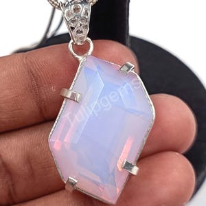 May include: A silver pendant with a large, faceted, light pink and white gemstone. The gemstone is set in a silver frame with four prongs and is suspended from a delicate silver chain. The pendant has the text "Tulipgems" on it.