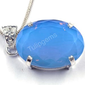 May include: A silver pendant necklace featuring a large, faceted, oval-shaped, light blue gemstone. The gemstone is set in a silver bezel and is suspended from a delicate silver chain. The text "Tulipgems" is visible on the gemstone.