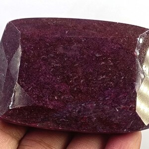Può includere: Una grande gemma rettangolare di colore magenta intenso con una superficie sfaccettata. La pietra presenta una finitura lucida e riflettente con inclusioni naturali visibili e un bordo distinto con molteplici sfaccettature.